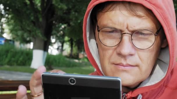 Un hombre con gafas se sienta en un banco en el parque y usa una tableta — Vídeos de Stock