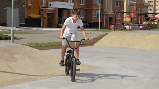 Un niño está montando trucos de ciclismo BMX en un parque de skate en un día soleado. Super cámara lenta — Vídeos de Stock