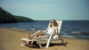 Beyaz bikinili genç kız yatıyor ve deniz kumlu plajda şezlong üzerinde sunbathes