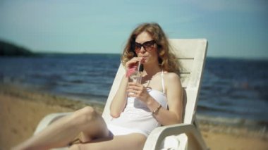 Beyaz bikinili genç bir kız yatıyor ve deniz kumlu sahilde bir tembel sunbathes ve bir kokteyl içer