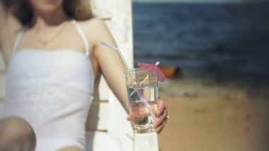 Beyaz bikinili genç bir kız yatıyor ve deniz kumlu sahilde bir tembel sunbathes ve bir kokteyl içer