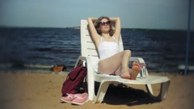 Beyaz bikinili genç kız yatıyor ve deniz kumlu plajda şezlong üzerinde sunbathes