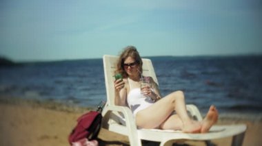 Beyaz bikinili genç bir kız yatıyor ve deniz kumlu sahilde bir tembel sunbathes ve akıllı telefon kullanır