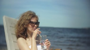 Beyaz bikinili genç bir kız yatıyor ve deniz kumlu sahilde bir tembel sunbathes ve bir kokteyl içer