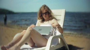 Beyaz bikinili genç bir kız yatıyor ve tans üzerinde bir deniz kumlu plajda şezlong ve bir dizüstü bilgisayar üzerinde çalışan
