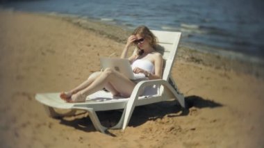 Beyaz bikinili genç bir kız yatıyor ve tans üzerinde bir deniz kumlu plajda şezlong ve bir dizüstü bilgisayar üzerinde çalışan