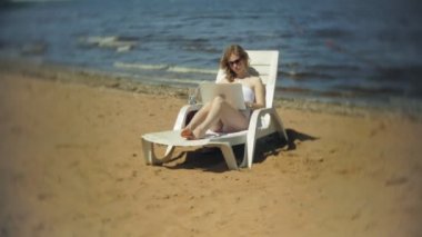 Beyaz bikinili genç bir kız yatıyor ve tans üzerinde bir deniz kumlu plajda şezlong ve bir dizüstü bilgisayar üzerinde çalışan