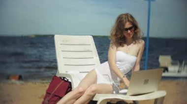 Beyaz bikinili genç bir kız yatıyor ve tans üzerinde bir deniz kumlu plajda şezlong ve bir dizüstü bilgisayar üzerinde çalışan