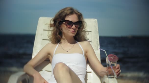 Ένα νεαρό κορίτσι σε ένα λευκό μπικίνι βρίσκεται και sunbathes σε μια ξαπλώστρα στην παραλία της θάλασσας αμμώδης και πίνει κοκτέιλ — Αρχείο Βίντεο