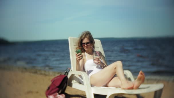 Ein junges Mädchen im weißen Bikini liegt und sonnt sich auf einer Liege am Sandstrand des Meeres und benutzt ein Smartphone — Stockvideo