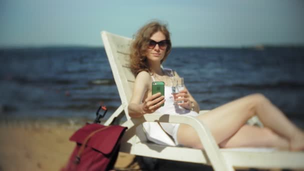 Молода дівчина в білому бікіні лежить і засмагає на шезлонгу на морському піщаному пляжі і використовує смартфон — стокове відео