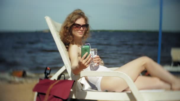 Mladá dívka v bílých bikinách leží a sunbathes na lehátkách na pláži u moře, písečná a používá smartphone — Stock video