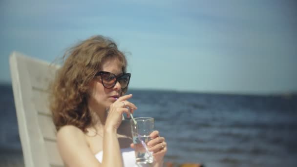 Una joven en bikini blanco se encuentra y toma el sol en una tumbona en la playa de arena marina y se bebe un cóctel. — Vídeo de stock