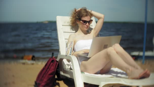 Ένα νεαρό κορίτσι σε ένα λευκό μπικίνι βρίσκεται και μαυρίσματα σε μια ξαπλώστρα στην αμμώδη παραλία και εργάζονται σε ένα φορητό υπολογιστή — Αρχείο Βίντεο