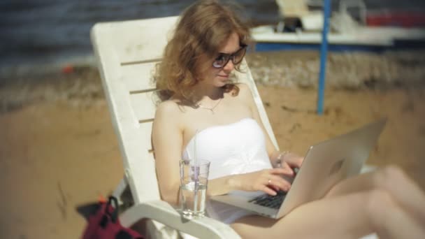 Ein junges Mädchen im weißen Bikini liegt und bräunt auf einem Liegestuhl am Sandstrand und arbeitet an einem Laptop — Stockvideo