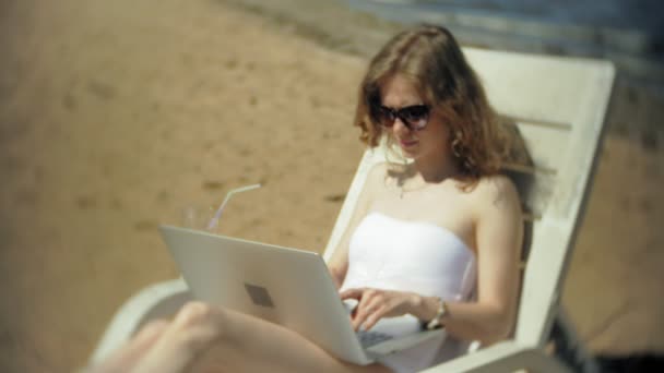 一个穿着白色比基尼的年轻女孩躺在海边沙滩上的躺椅上上晒黑, 正在用笔记本电脑 — 图库视频影像
