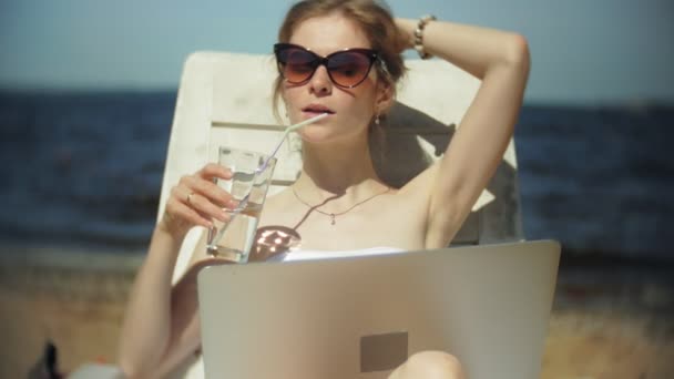 Una joven en un bikini blanco se encuentra y broncea en una tumbona en una playa de arena de mar y está trabajando en un ordenador portátil — Vídeos de Stock