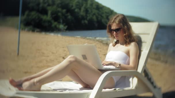 Молода дівчина в білому бікіні лежить і пил на шезлонгу на морському піщаному пляжі і працює на ноутбуці — стокове відео
