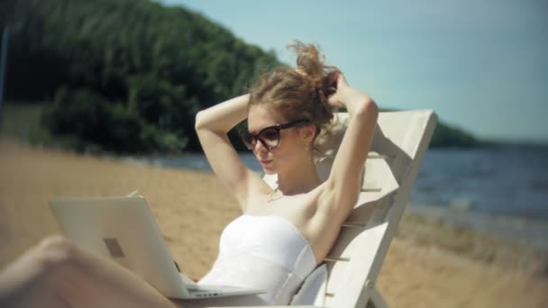 En ung flicka i en vit bikini ligger och tans på en solstol på en sandig strand och arbetar på en bärbar dator — Stockvideo