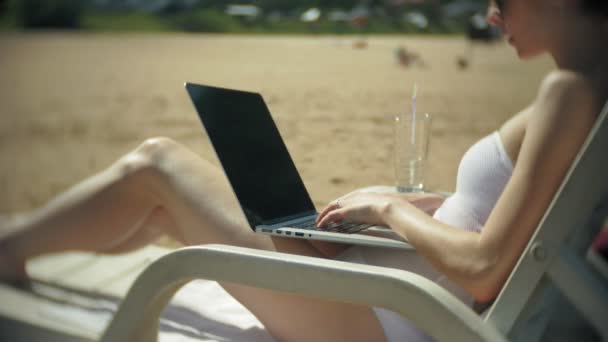 Una giovane ragazza in bikini bianco si sdraia e abbronza su una sdraio su una spiaggia di sabbia marina e sta lavorando su un computer portatile — Video Stock