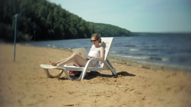 Egy fiatal lány, egy fehér bikiniben fekszik és varádics a nyugágyon a tenger homokos strandján és működő-ra egy laptop — Stock videók