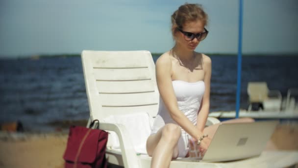 Una giovane ragazza in bikini bianco si sdraia e abbronza su una sdraio su una spiaggia di sabbia marina e sta lavorando su un computer portatile — Video Stock