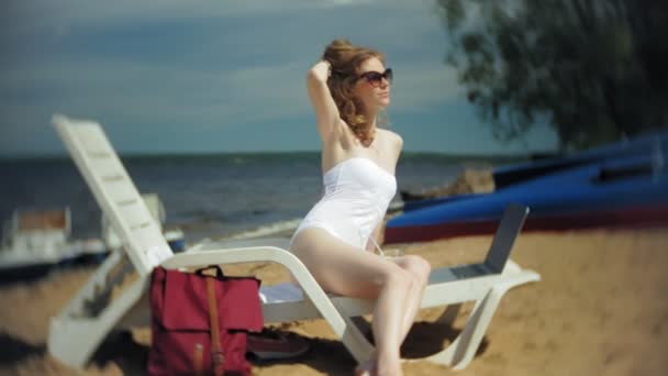 Beyaz bikinili genç bir kız yatıyor ve tans üzerinde bir deniz kumlu plajda şezlong ve bir dizüstü bilgisayar üzerinde çalışan — Stok video