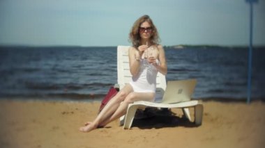 Beyaz bikinili genç bir kız yatıyor ve tans üzerinde bir deniz kumlu plajda şezlong ve bir dizüstü bilgisayar üzerinde çalışan