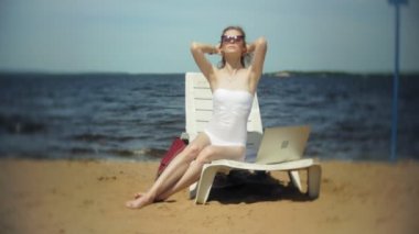 Beyaz bikinili genç bir kız yatıyor ve tans üzerinde bir deniz kumlu plajda şezlong ve bir dizüstü bilgisayar 4 k çalışma