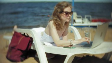 Beyaz bikinili genç bir kız yatıyor ve tans üzerinde bir deniz kumlu plajda şezlong ve bir dizüstü bilgisayar üzerinde çalışan