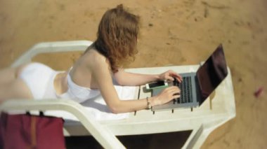 Beyaz bikinili genç bir kız yatıyor ve tans üzerinde bir deniz kumlu plajda şezlong ve bir dizüstü bilgisayar üzerinde çalışan