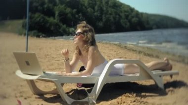 Beyaz bikinili genç bir kız yatıyor ve tans üzerinde bir deniz kumlu plajda şezlong ve bir dizüstü bilgisayar üzerinde çalışan