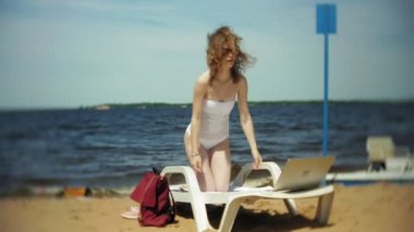 Beyaz bikinili genç bir kız yatıyor ve tans üzerinde bir deniz kumlu plajda şezlong ve bir dizüstü bilgisayar üzerinde çalışan
