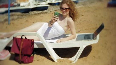 Beyaz bikinili genç bir kız yatıyor ve tans üzerinde bir deniz kumlu plajda şezlong ve bir dizüstü bilgisayar üzerinde çalışan