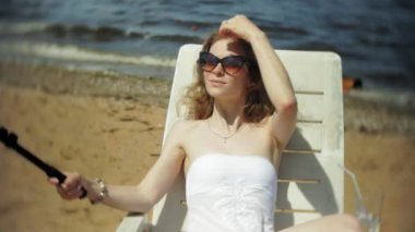 Beyaz bikinili genç bir kız yatıyor ve bir deniz kumlu plajda şezlong ve fotoğrafları Smartphone cep telefonu ile Selfi sunbathes