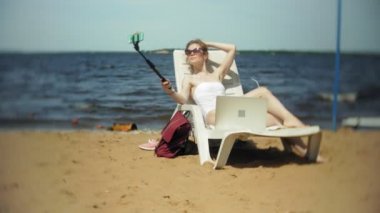 Beyaz bikinili genç bir kız yatıyor ve bir deniz kumlu plajda şezlong ve fotoğrafları Smartphone cep telefonu ile Selfi sunbathes