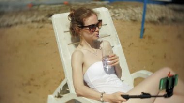 Beyaz bikinili genç bir kız yatıyor ve bir deniz kumlu plajda şezlong ve fotoğrafları Smartphone cep telefonu ile Selfi sunbathes