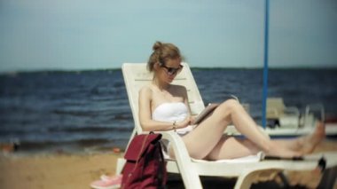 Beyaz bikinili genç bir kız yatıyor ve tans üzerinde bir deniz kumlu plajda şezlong ve bir tablet üzerinde çalışan