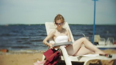 Beyaz bikinili genç bir kız yatıyor ve tans üzerinde bir deniz kumlu plajda şezlong ve bir dizüstü bilgisayar üzerinde çalışan