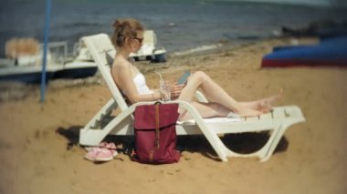 Beyaz bikinili genç bir kız yatıyor ve tans üzerinde bir deniz kumlu plajda şezlong ve bir tablet üzerinde çalışan