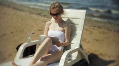 Beyaz bikinili genç bir kız yatıyor ve tans üzerinde bir deniz kumlu plajda şezlong ve bir tablet üzerinde çalışan