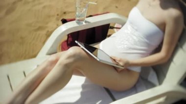 Beyaz bikinili genç bir kız yatıyor ve tans üzerinde bir deniz kumlu plajda şezlong ve bir tablet üzerinde çalışan