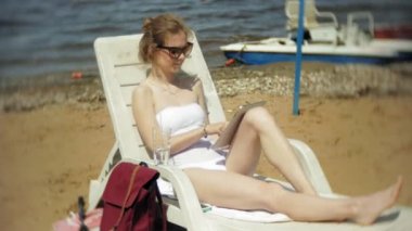 Beyaz bikinili genç bir kız yatıyor ve tans üzerinde bir deniz kumlu plajda şezlong ve bir tablet üzerinde çalışan