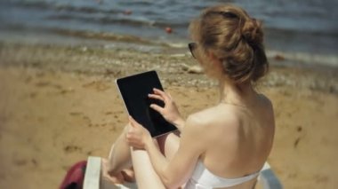Beyaz bikinili genç bir kız yatıyor ve tans üzerinde bir deniz kumlu plajda şezlong ve bir tablet üzerinde çalışan