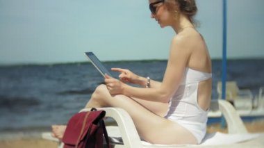 Beyaz bikinili genç bir kız yatıyor ve tans üzerinde bir deniz kumlu plajda şezlong ve bir tablet üzerinde çalışan