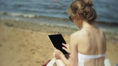 Beyaz bikinili genç bir kız yatıyor ve tans üzerinde bir deniz kumlu plajda şezlong ve bir tablet üzerinde çalışan