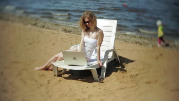 En ung flicka i en vit bikini ligger och tans på en solstol på en sandig strand och arbetar på en bärbar dator — Stockvideo