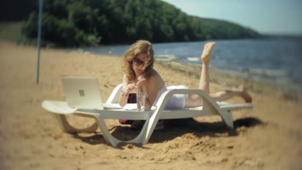 Egy fiatal lány, egy fehér bikiniben fekszik és varádics a nyugágyon a tenger homokos strandján és működő-ra egy laptop — Stock videók
