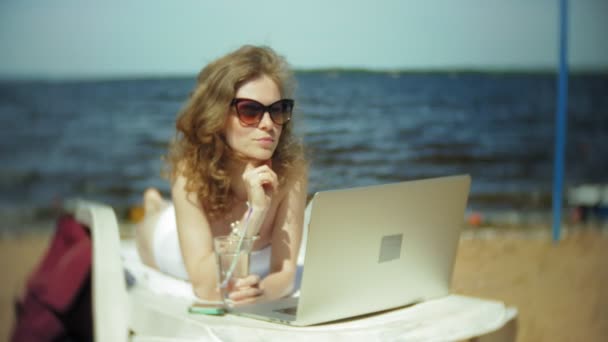 Ein junges Mädchen im weißen Bikini liegt und bräunt auf einem Liegestuhl am Sandstrand und arbeitet an einem Laptop — Stockvideo