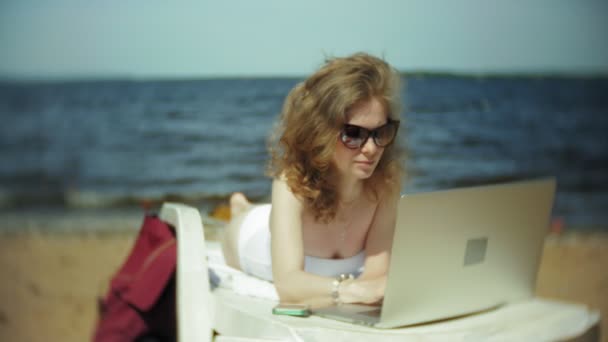 一个穿着白色比基尼的年轻女孩躺在海边沙滩上的躺椅上上晒黑, 正在用笔记本电脑 — 图库视频影像